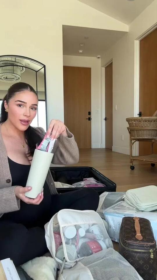 Olivia Culpo sitting on the floor packing a luxury hospital bag, holding a container of wellness items with open suitcases and beauty products around her.