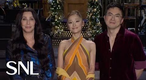 Cher, Bowen Yang, and Simu Liu standing together on the Saturday Night Live stage in front of holiday decorations, smiling during a promo for the Christmas 2025 episode.