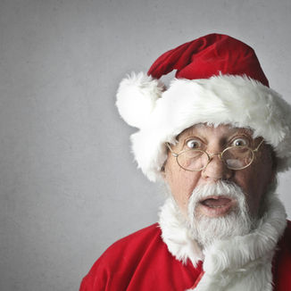 Surprised man dressed as Santa Claus with wide eyes and open mouth, wearing a red suit, white beard, and round glasses, standing against a plain gray background.