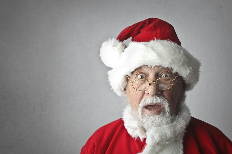Surprised man dressed as Santa Claus with wide eyes and open mouth, wearing a red suit, white beard, and round glasses, standing against a plain gray background.