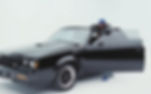Kendrick Lamar in a blue cap and leather jacket leaning on the open door of a black Buick GNX against a minimalist white background.