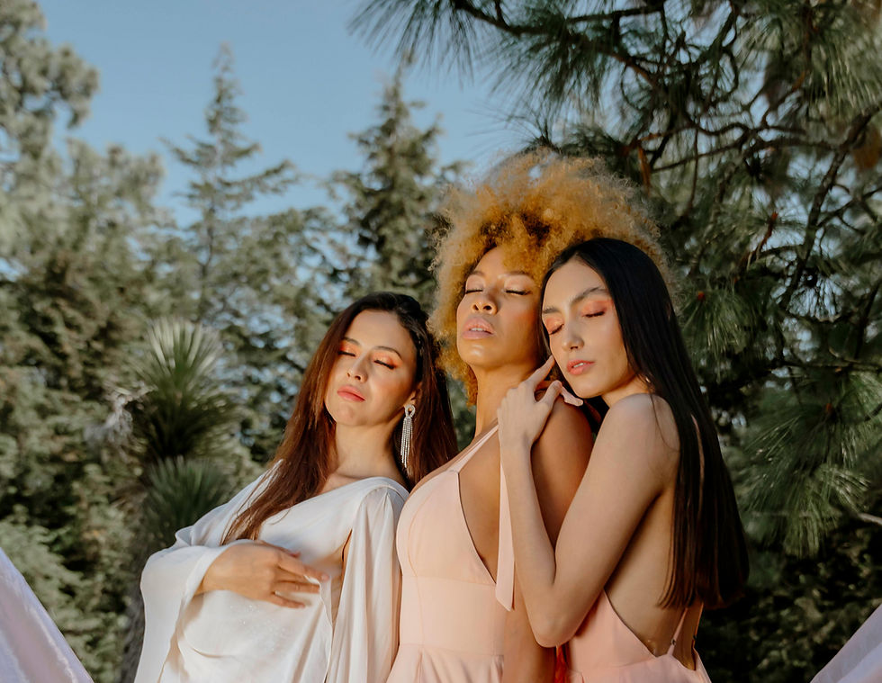 Three women in flowing blush and ivory gowns pose outdoors among tall trees, creating an ethereal fashion editorial scene.