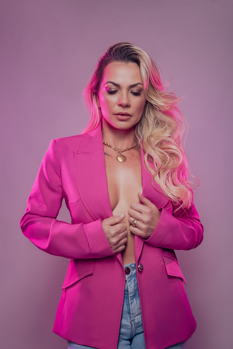 Gisely Garcia poses against a soft mauve studio backdrop wearing a vibrant pink blazer, light-wash jeans, and gold jewelry, with long blonde waves styled over one shoulder.