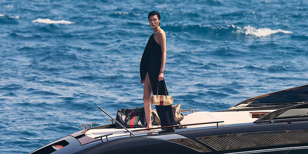 Gucci campaign image showing a model in a black one-shoulder outfit standing on a yacht at sea while holding a small patterned handbag.