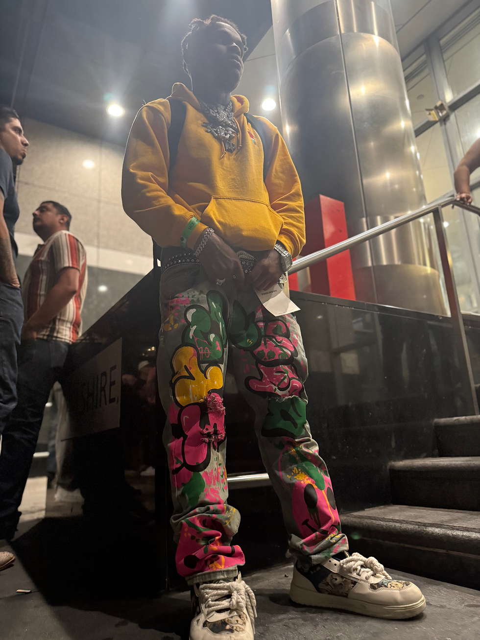 Young Drew wearing a yellow hoodie, colorful graphic pants, white sneakers, a backpack, and layered jewelry while standing near an indoor staircase with other people nearby.
