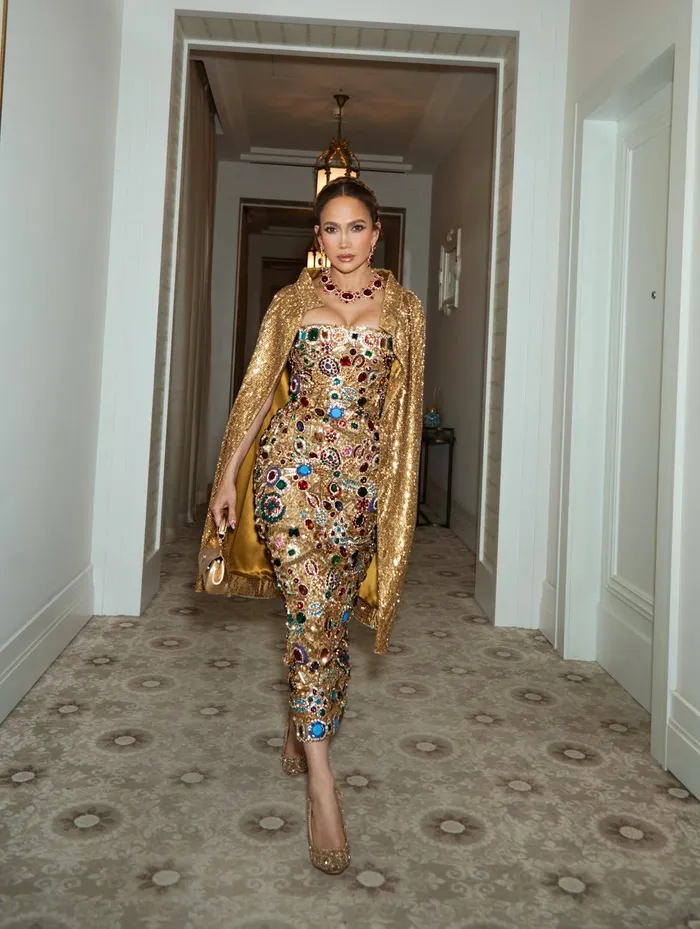 Jennifer Lopez wearing a gold, jewel-covered evening gown with a shimmering cape in a luxury hotel hallway.
