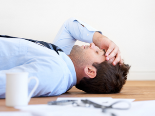 Business owner in blue shirt and tie lying on wooden floor, hand on forehead, looking exhausted, burned out. Journey Consulting