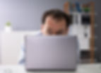 Business owner hides behind laptop, avoiding difficult conversations with employees. Blurred bookshelves in background.