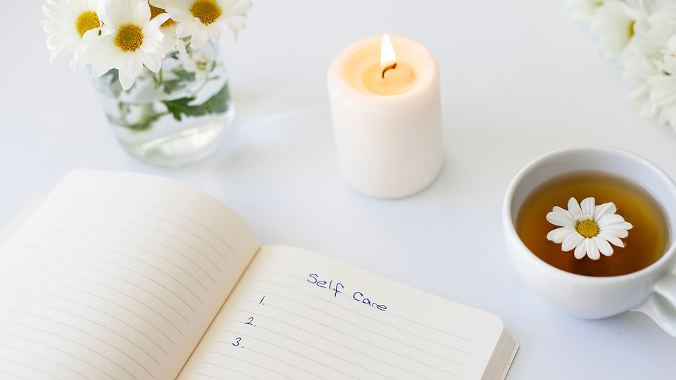 Open notebook labeled "Self care," candle, daisies in vase, and a cup of tea with flower on a white surface. Calm and serene setting.