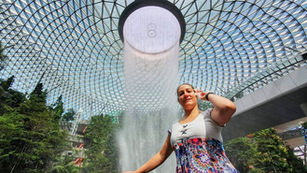 Christian Money Mindset coach - Woman poses joyfully under a large indoor waterfall. Glass dome ceiling with geometric patterns and lush greenery surrounds her.