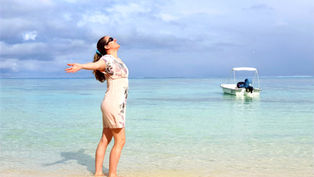 Woman in floral dress stands on a beach with arms outstretched, gazing upwards. Boat floats on clear water. Sky is cloudy.