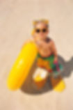 A boy on the beach in snorkel flippers, snorkel mask, sunglasses carrying a sand bucket and floatie