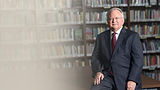Man in a suit sitting in front of a bookshelf.