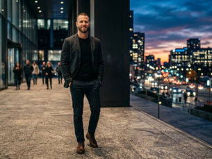 A confident bearded man smiling while walking through a sleek, modern urban plaza at sunset. He is wearing a dark casual blazer, black t-shirt, fitted pants, and brown leather boots. In the background, city traffic lights and a dramatic sunset sky create a powerful atmosphere, perfectly capturing the core concept of modern masculinity for Imperium Max V.