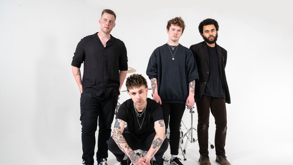 Four Band Members Dressed In Black Posing At Different Heights In Front Of A Drum Kit For Album Promotion.