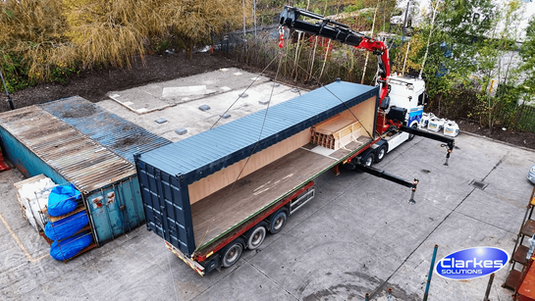 A sipping container being lifted off the back of a flat bed lorry by a crane.