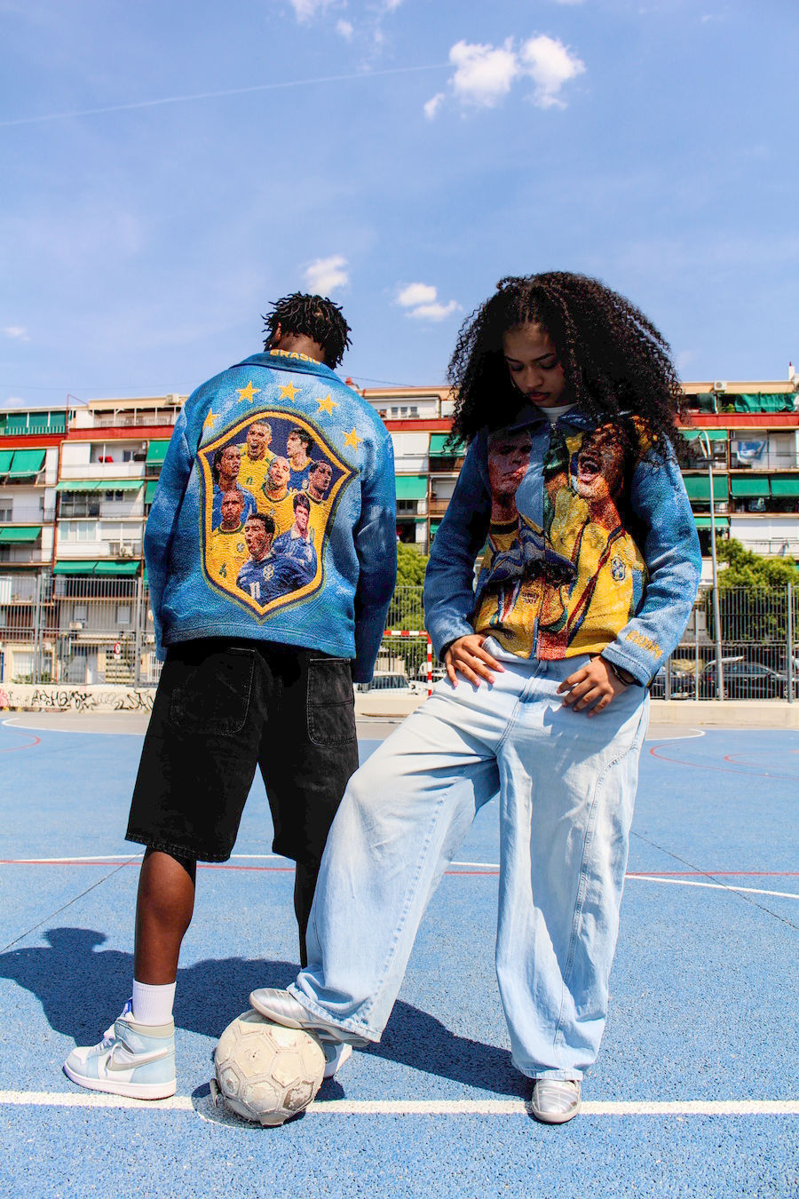 Two models wearing the Joga Bonito Tapestry Jacket on an outdoor football court, showcasing the front and back design.