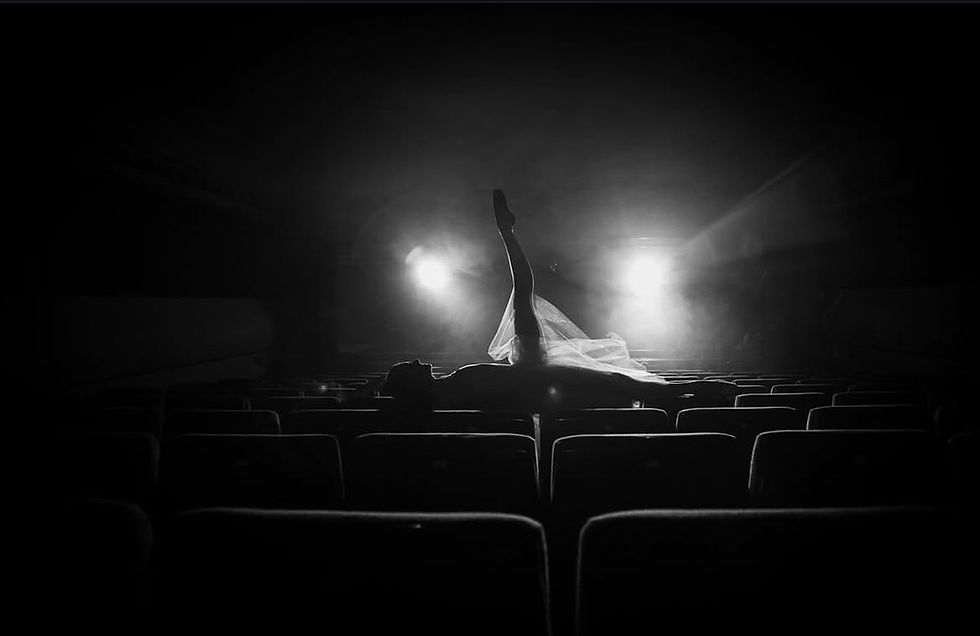 Black and white artistic photo of a dancer lying across theater seats with one leg extended upward