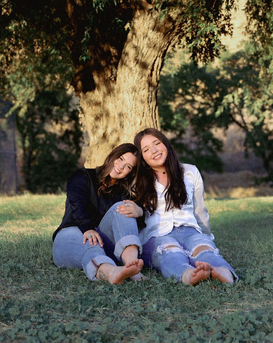 Two friends sitting on grass in front of a tree; one with her head on the others shoulder