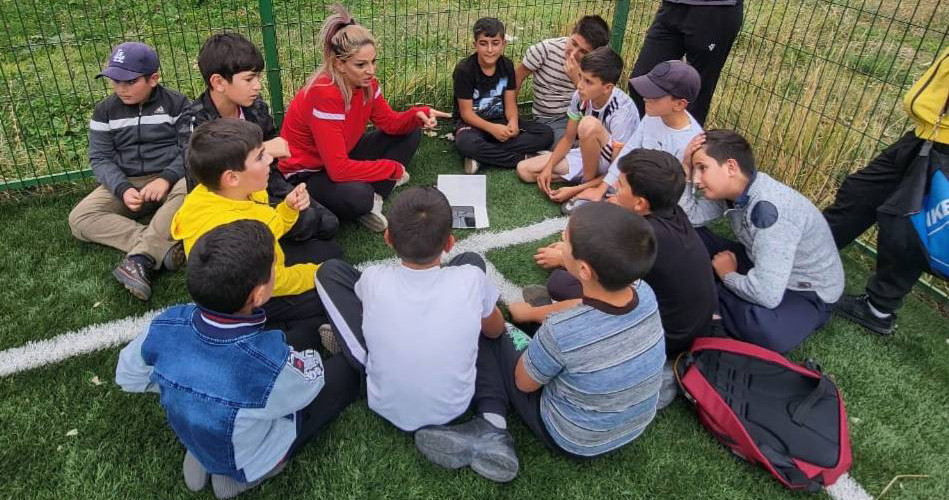 A group of soccer players sit in a circle on the ground with their coach