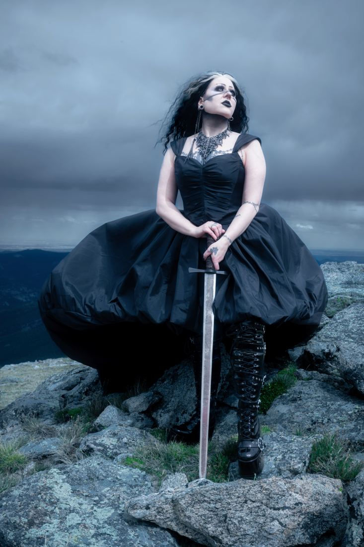 Woman seated on rocks with a sword planted in front of her, storm clouds behind