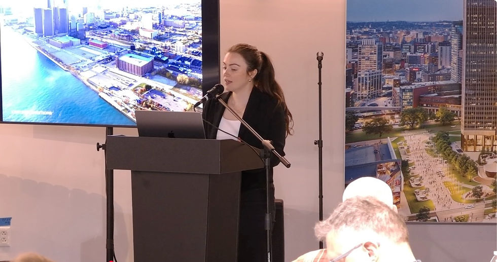 Presenter beside an aerial Renaissance Center slide and a conceptual riverfront development display.