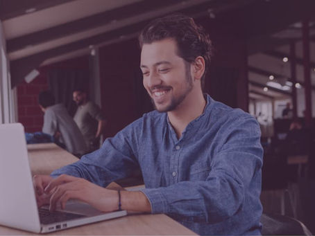 A smiling man in a blue linen shirt works on a laptop in an open, modern workspace with other people visible in the background.