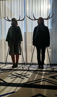 Olga Zitluhina and Fiona Millward standing below deer antlers, framed with a window behind them
