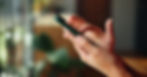 Hands holding a smartphone, typing. Blurred indoor background with soft light, green and beige tones. Calm and focused mood.