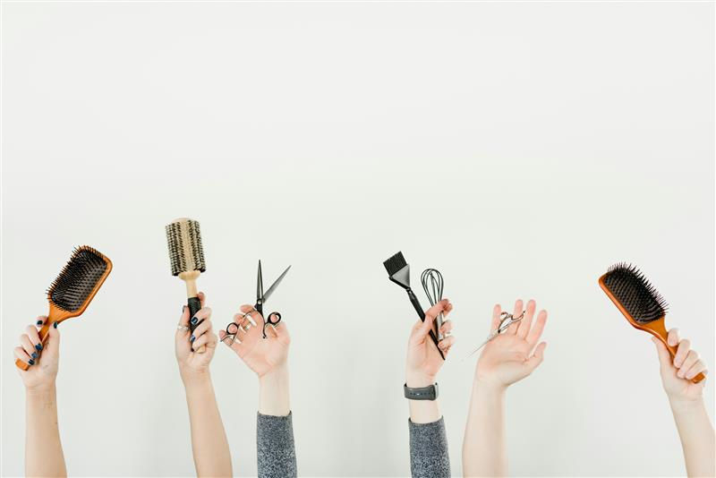 Hands hold various hair styling tools against a white background: brushes, scissors, combs. The mood is creative and professional.