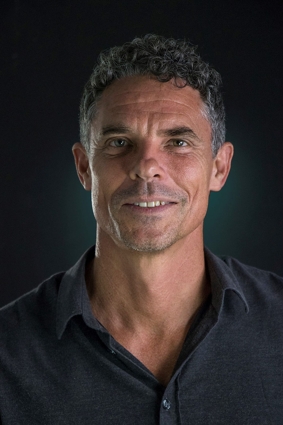 Smiling man with curly hair in a dark shirt, set against a black background. The mood is positive and confident.