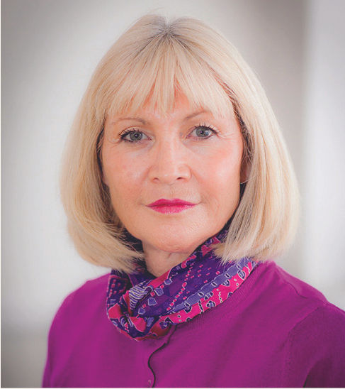 A woman with short blonde hair wears a bright purple top and a patterned scarf, looking directly at the camera with a neutral expression.