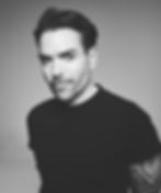 Black-and-white portrait of a man in a black T-shirt with a tattooed arm, posing against a plain gray background with a slight smile