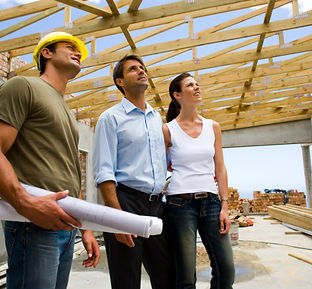 Contractor holding architectural drawings while speaking with man and woman, all looking up at rafters being installed on a a