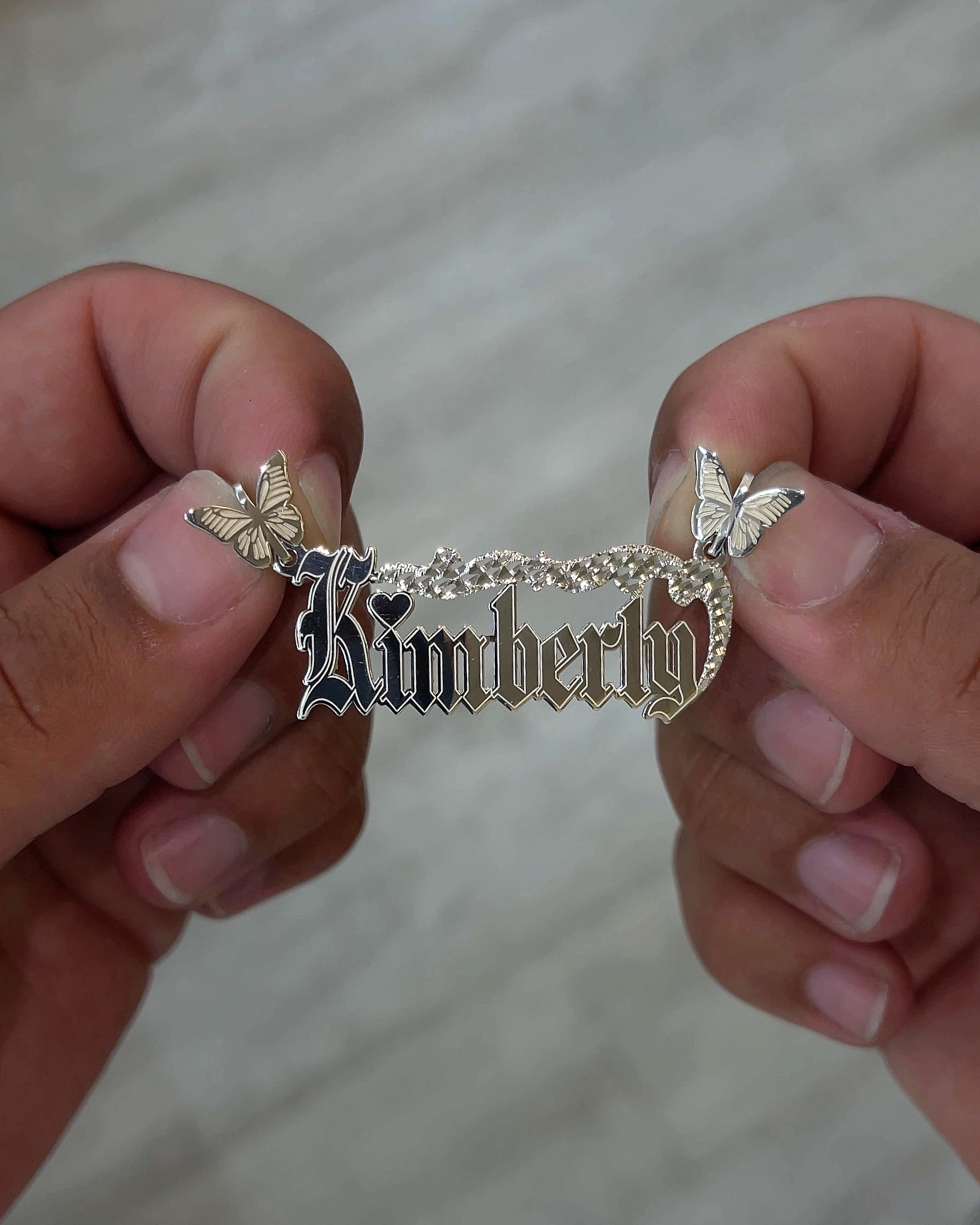 Hands holding sterling silver nameplate pendant in old english font with butterflies as the bails and diamond cut texture