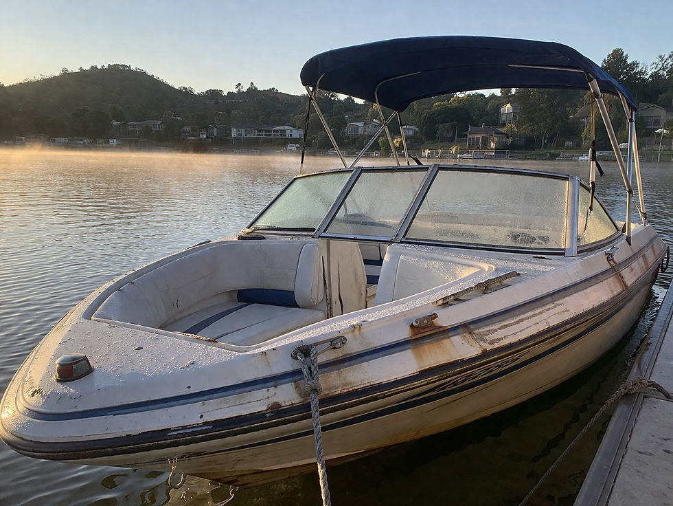 Weathered white and blue motorboat docked on a calm misty lake at sunrise, with hillside houses in the background