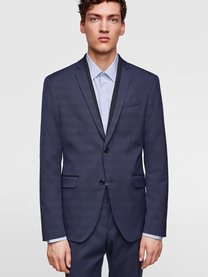 Man in dark navy blue suit jacket and pants, wearing a light blue shirt with a black collar, standing against a white background.