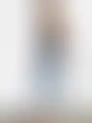 Woman wearing black top and jeans posing against white wall Charme