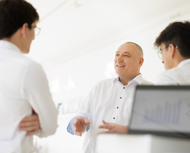 eine Besprechungssituation mit 3 Personen. Die Person in der Mitte erklärt etwas und die beiden Zuhörer sieht man von hinten