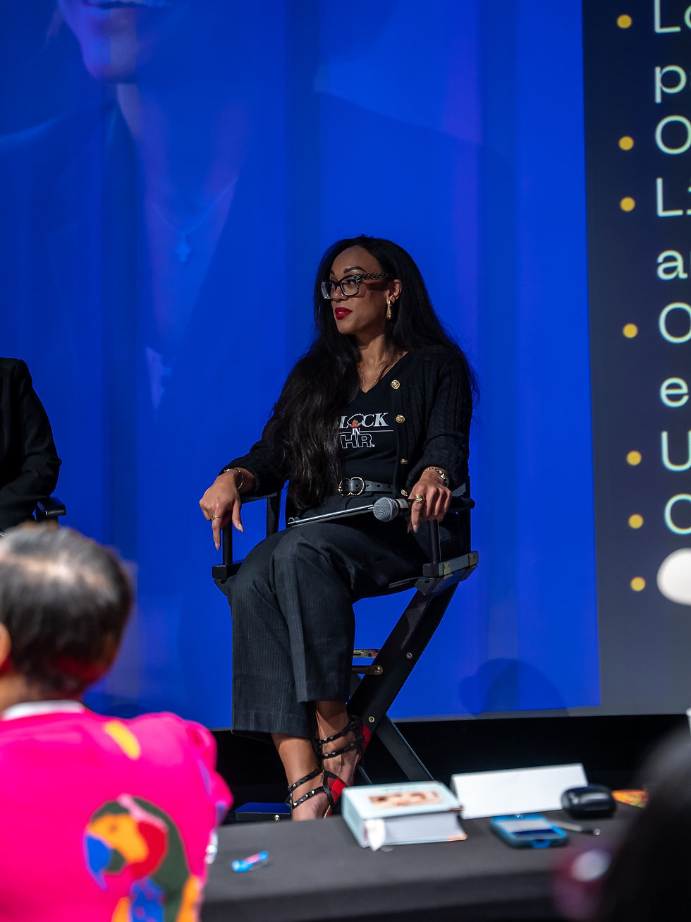 Woman in glasses seated on stage, with text visible, The Black in HR
