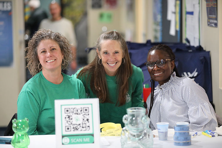 Three diverse women smile together. Two wear Froggy's Closet green tshirts. They are at a school charity event for kids, the background is blurred