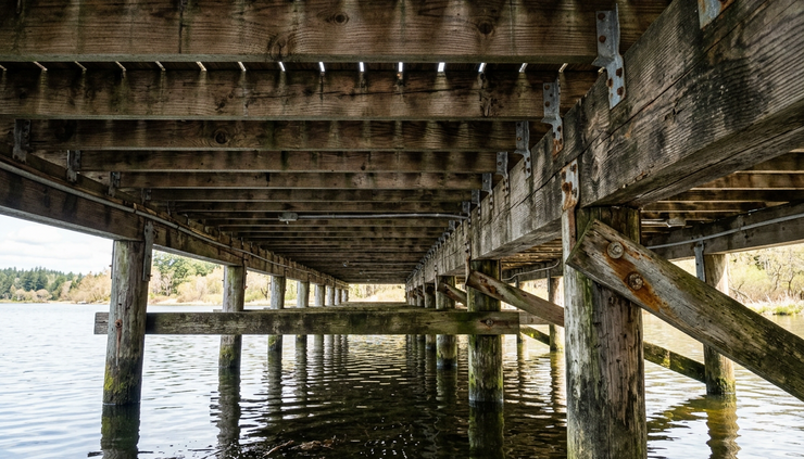 Low-angle view beneath a dock showing beams, joists, hardware, and water below