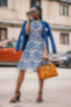 A woman standing in a street (with a building and two parked cars behind her), wearing blue, heart-shaped sunglasses; a cobalt blue, tartan patterned blazer over her shoulders; a royal blue, black and white patterned a-line dress and black, ankle strap stiletto heels. The woman has long braided hair tied into a ponytail and is holding a rustic tan coloured kelly handbag.
