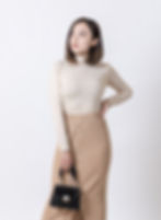 A woman looking away and posing, wearing a high quality cream top, tan woollen skirt and holding a black handbag
