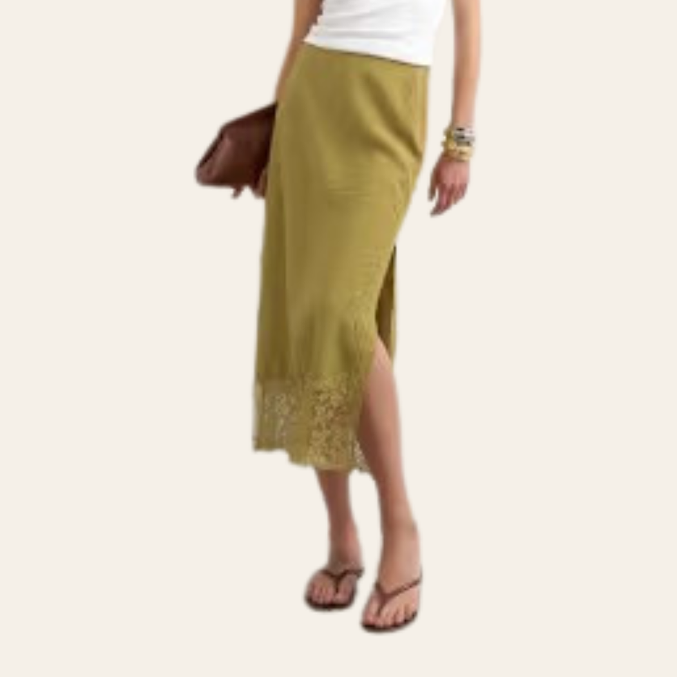 Woman in a white top and olive skirt with lace hem, holding a brown clutch, posing in a cream background. Belonging to a capsule wardrobe for a woman in her 30s.