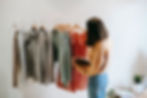 A woman with shoulder length brunette hair, holding a notebook and wearing a yellow ochre coloured jumper and light blue jeans has her face away from the camera and is examining a free standing clothing rail, set against a white backdrop. The rail has the following clothes hanging on wooden hangers from it: a beige ribbed jumper; three marl grey shirts; three mint green cropped cardigans and three orange t-shirts.