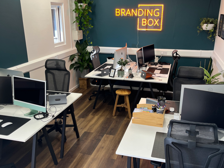 Modern office space with desks, computers, and plants. Neon sign reads "BRANDING BOX" on dark teal wall. Warm lighting and wooden floor.
