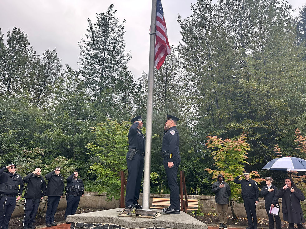 Juneau Police Department Deputy Chief Krag Campbell and Detective Kirt Stage-Harvey salute after raising the U.S. flag to half mast during a ceremony Thursday, Sept. 11, 2024, commemorating the 24th anniversary of the 9/11 terrorist attacks. At right, Sara Radke Brown sings the national anthem for the more than 100 people attending the event at Riverside Rotary Park. (Mark Sabbatini / Juneau Independent)