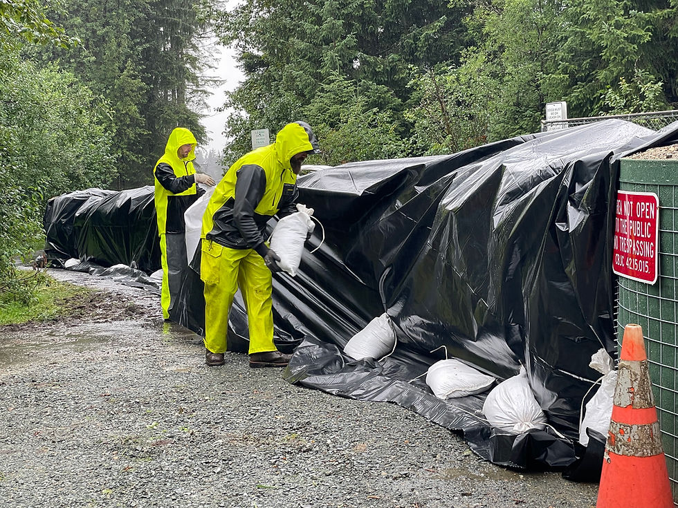 City workers deposit sandbags to hold in place heavy plastic sheeting wrapped around supersacks at the east end of now-closed Mendenhall River pedestrian bridge on Monday, Aug. 11, 2025. Their work filled in the gap on the trail between rows of HESCO barriers. (Laurie Craig / Juneau Independent)