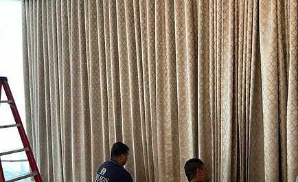 Two men are collaborating on installing a curtain in a well-lit room, focused on their task with tools in hand.